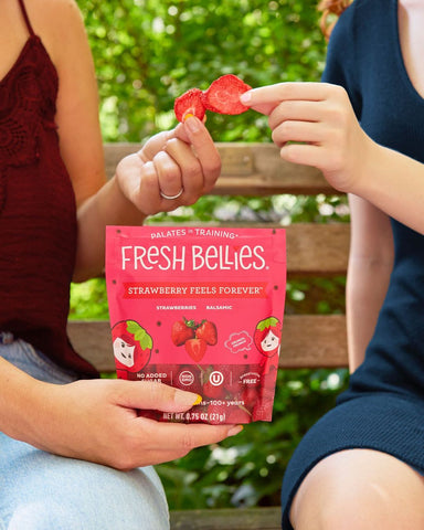 Close-up of two people's hands, each of whom is holding a Fresh Bellies strawberry snack. One woman holds the bag of snacks in her other hand