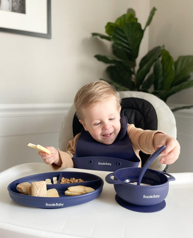 Baby in a high chair reaching for a spoon inside a bowl. Baby's bib, bowl, spoon, and plate on the high chair are all Bazzle Baby brand