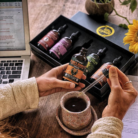 hands dropping an extract from a bottle into a cup of coffee. 4 more bottles in a gift set behind the coffee