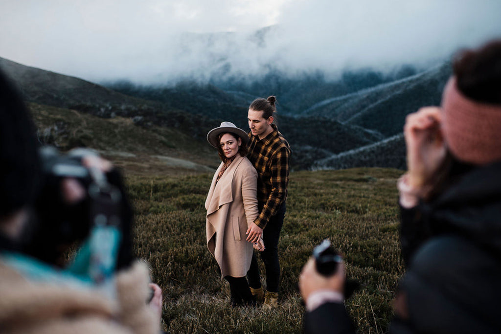 Male and female couple pose in front of a misty valley whilst group of photographers capture their image