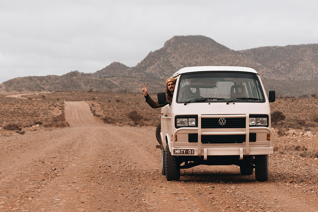 Male driver wearing Levi Mustard beanie throwing a hand peace sign out the window of vintage van parked on the side of a dirt road