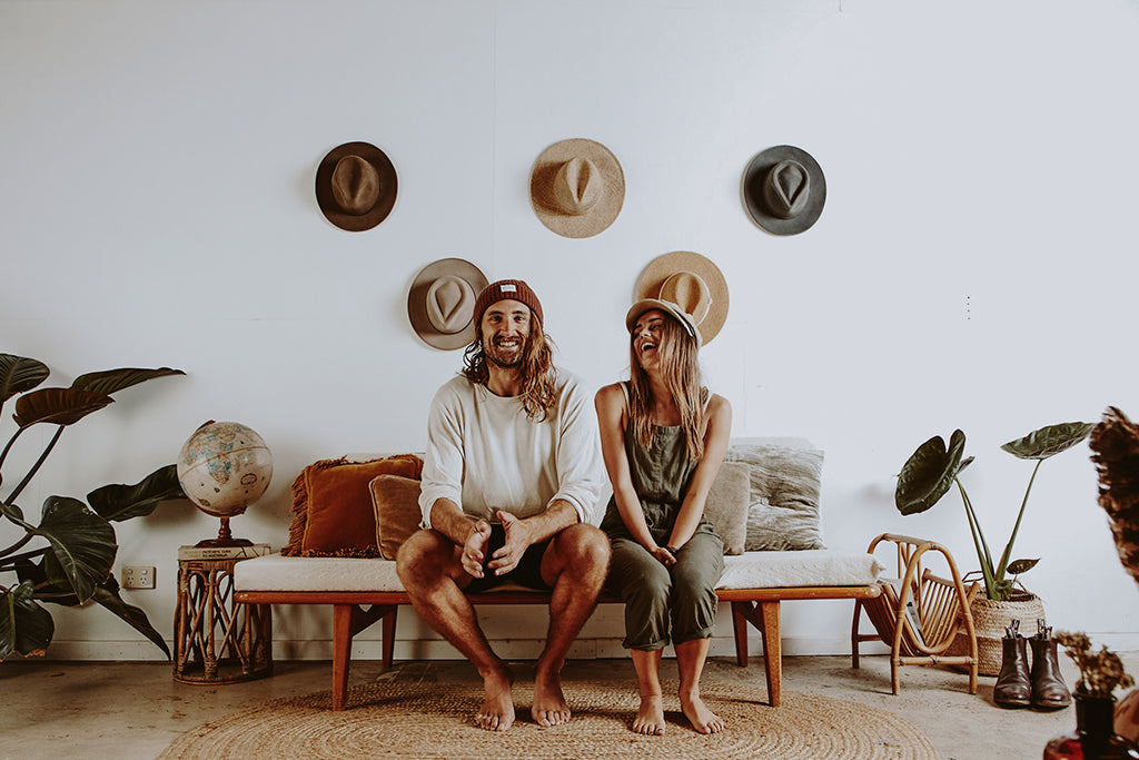 Owners of Will & Bear Alex and Lauren sit side by side in front of their collection of hanging wide brim hats