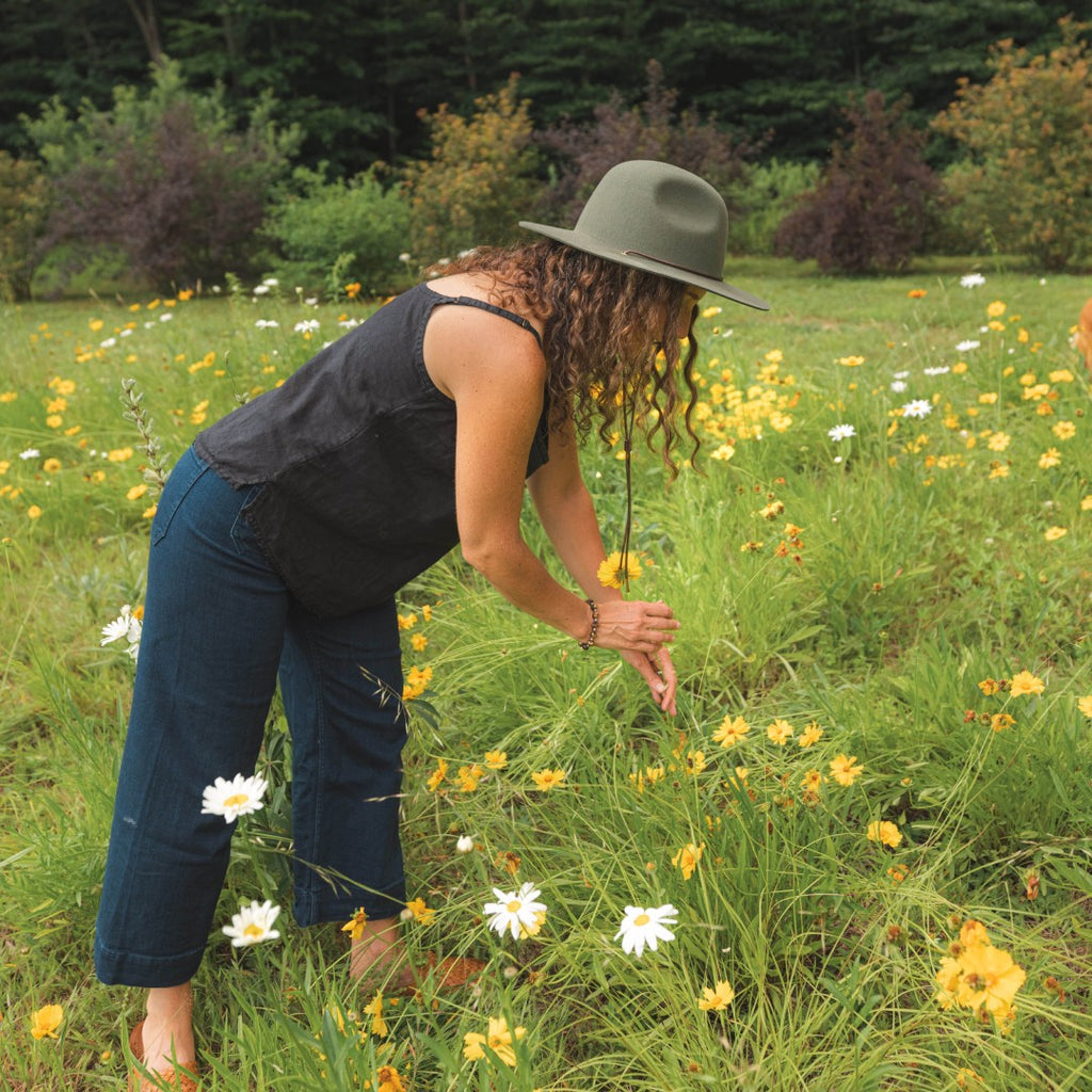 A woman with long curly hair wearing a wide brim sun hat is picking flowers in a grassy field