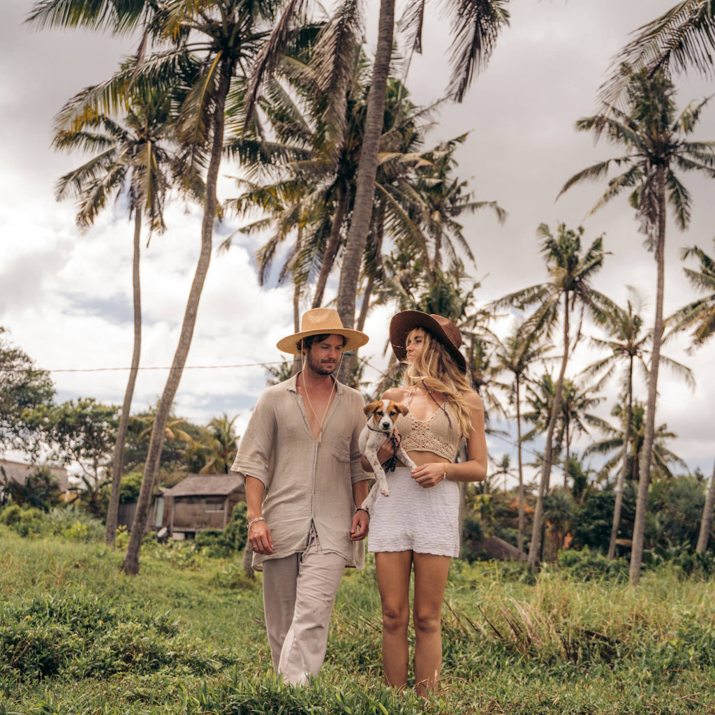 Man and woman wearing wide brim sun hats holding a puppy while walking through grass and palm trees