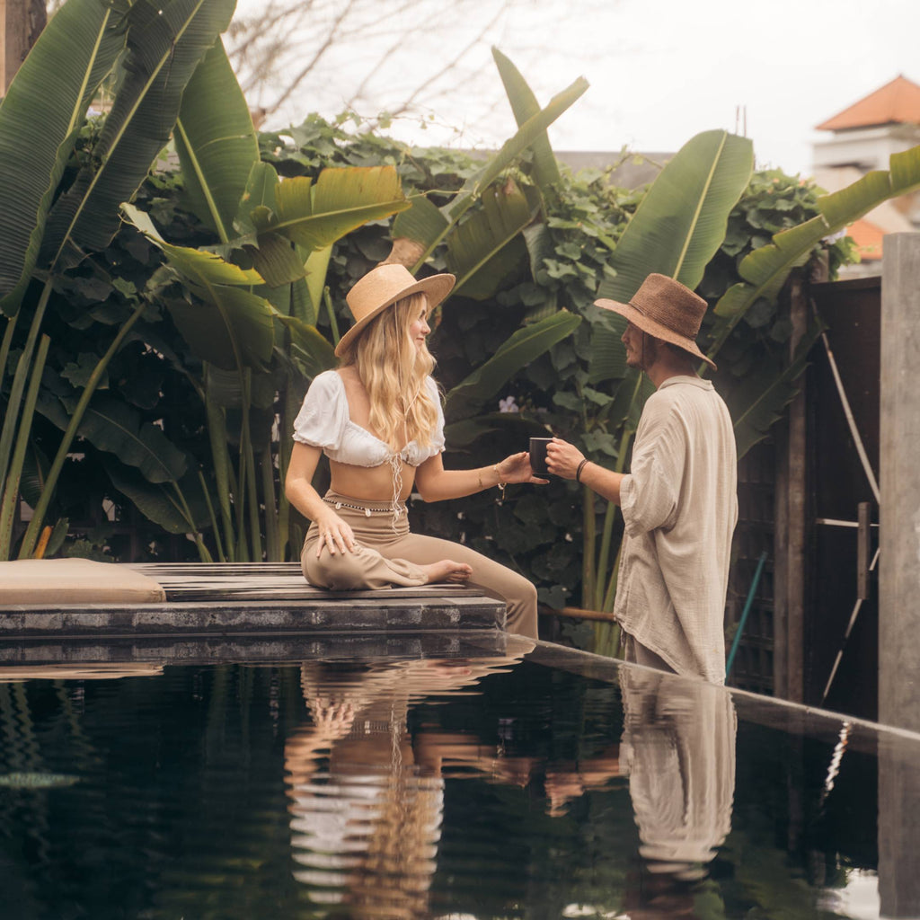 A man with short hair wearing a wide brim straw hat is handing a cup of coffee to a woman with long blonde hair wearing a wide brim sun hat