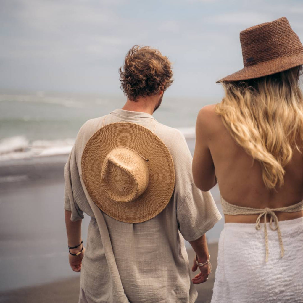 A man with short hair wearing a wide brim sun hat on his back is walking along the beach