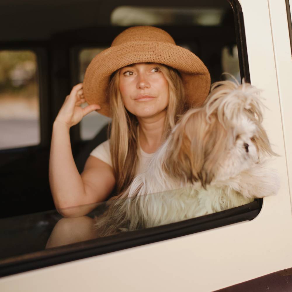 Woman with long brown hair wearing a wide brim straw hat on a road trip with her dog