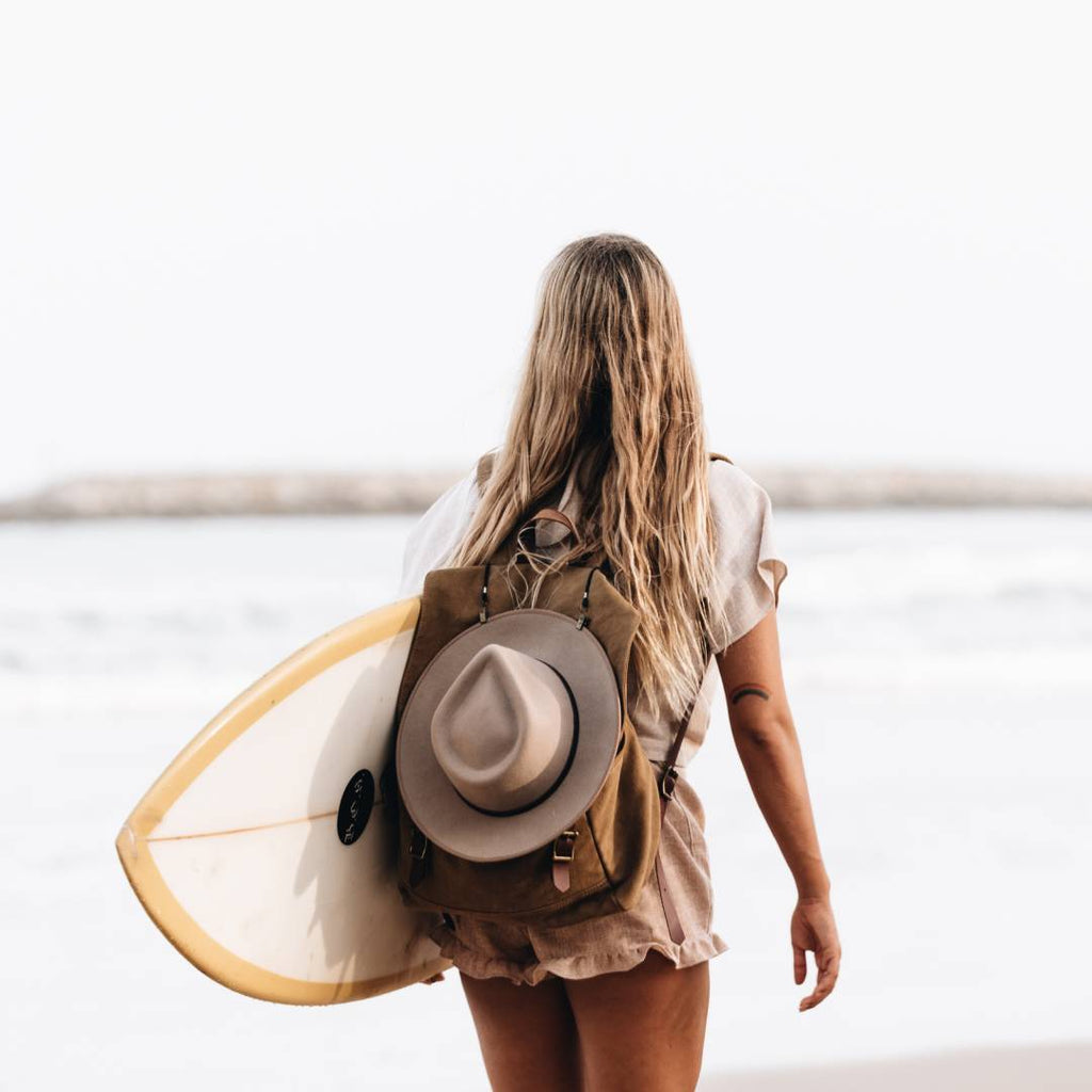 Woman with long blonde hair carrying a surf board with a wool hat strapped to her backpack