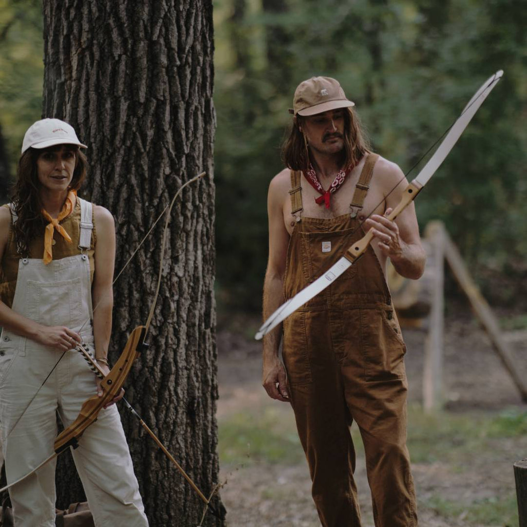 A man and a woman wearing caps, doing archery at a summer camp