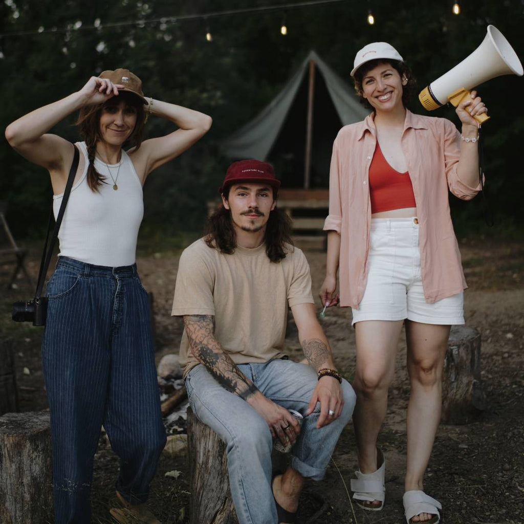 Three adults sitting at a campsite wearing caps, one is holding a megaphone