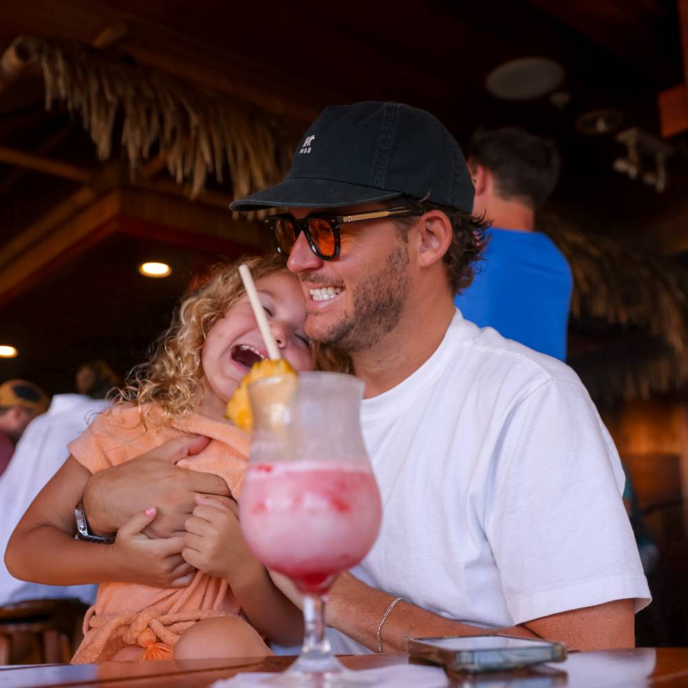 Man with short curly hair, wearing a black cap is drinking a cocktail in hawaii