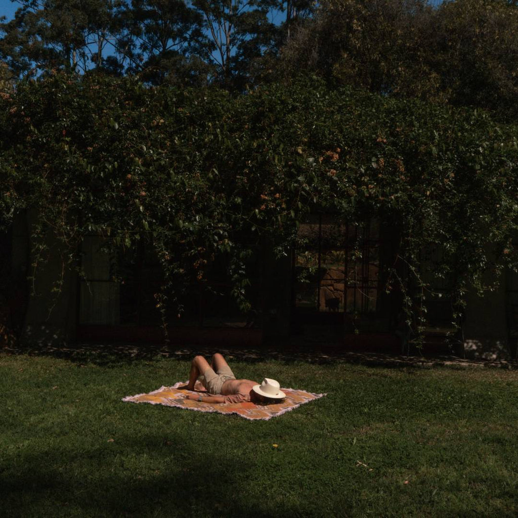 A man with long brown hair is sunbathing in a backyard with a wide brim straw hat covering his face