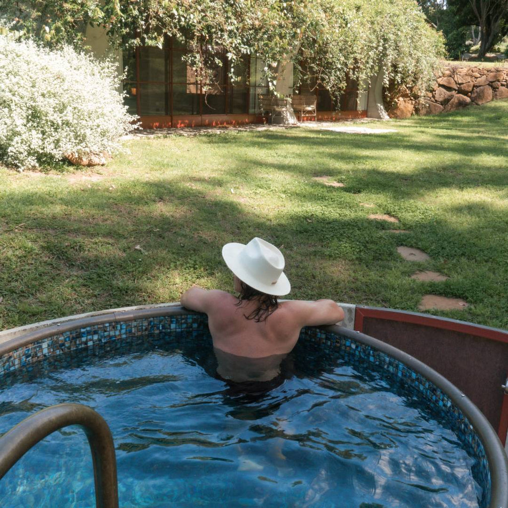 A man with long dark brown hair wears a straw wide brim hat while swimming in a small backyard pool