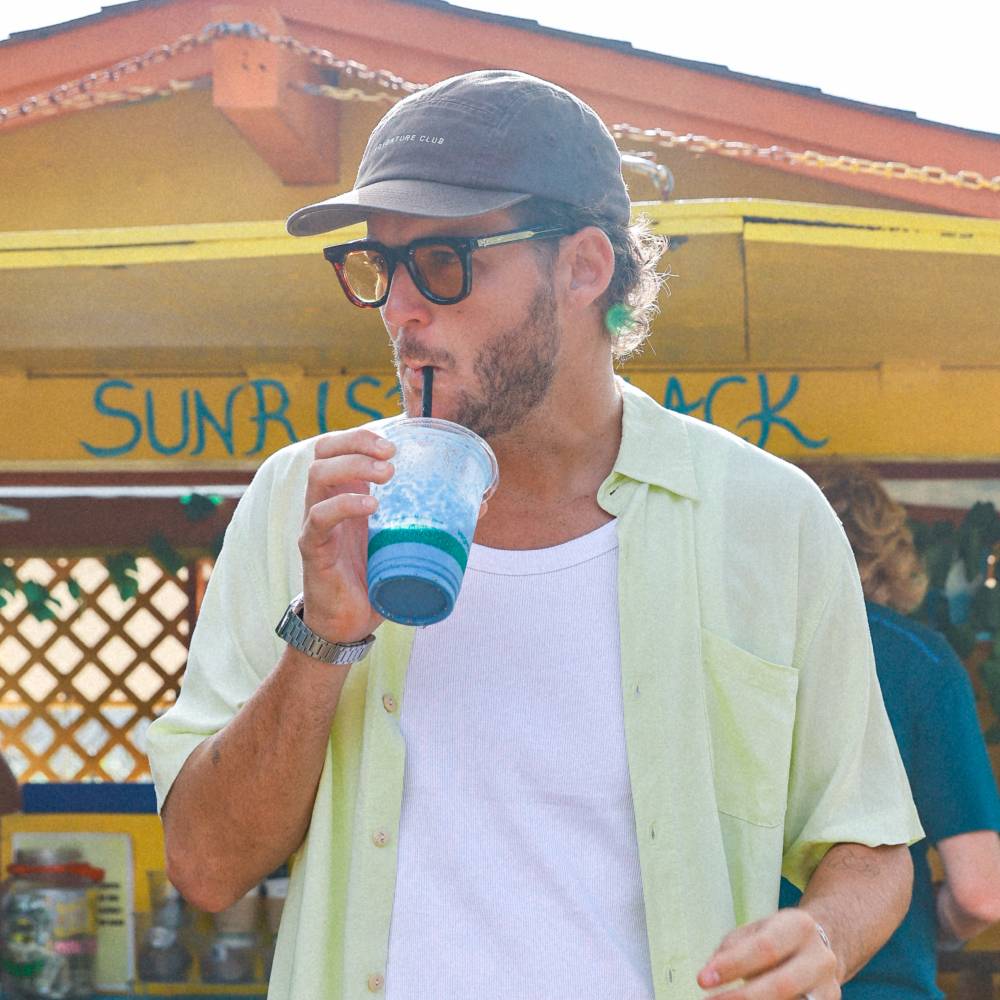Man with short curly hair wearing a brown cap while drinking a smoothie outside Sunrise Shack in Hawaii