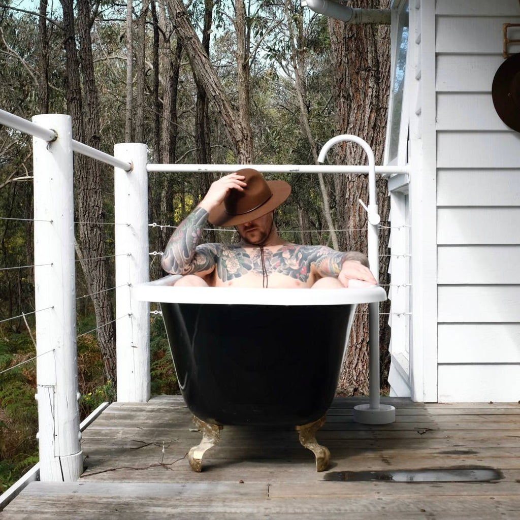 A man with short hair is wearing a wide brim wool hat while sitting in an outdoor bath in the Australian bush