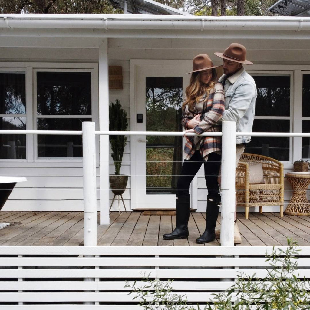 Young adult couple wearing wide brim wool hats, hugging and standing on the deck of an Australian beach shack