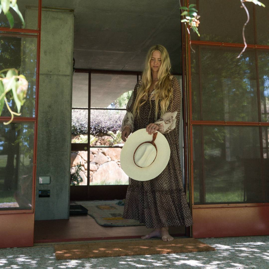 A woman with long blonde hair is holding a cowboy hat and standing in the middle of a doorway leading to the outside