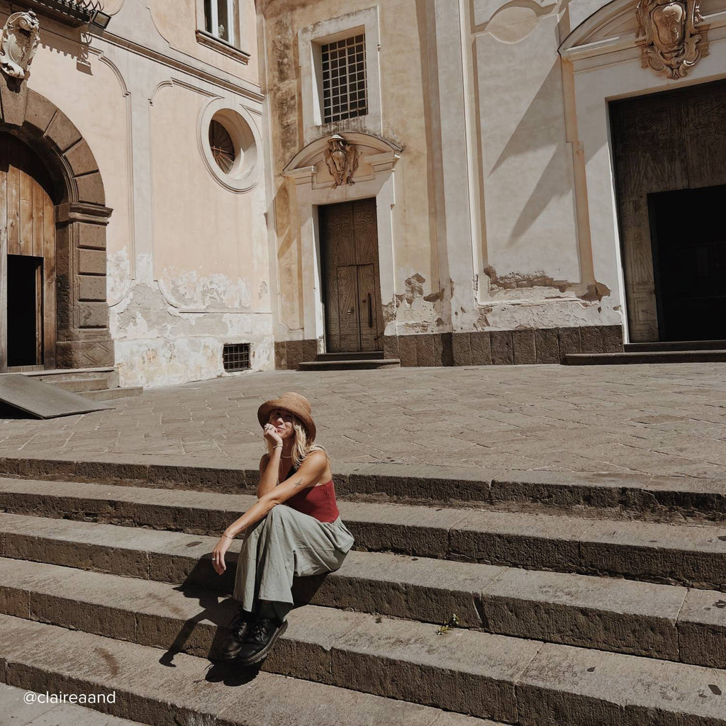 A young woman with long blonde hair wearing a straw bucket hat sits on the steps of an old European building