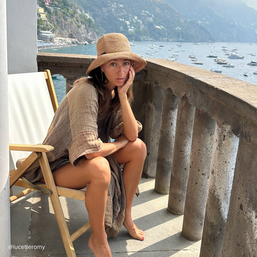 A young woman with shoulder length, wearing a straw bucket hat is sitting on a balcony in the mediterranean