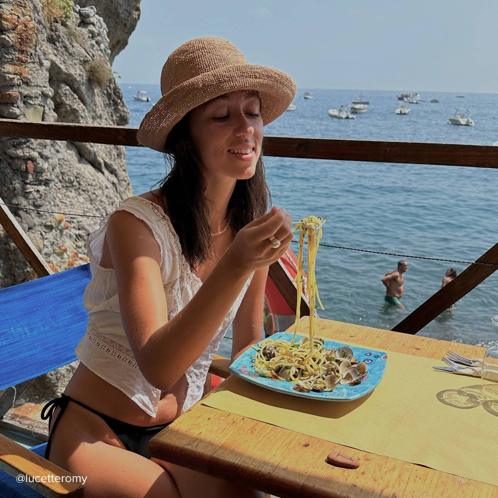 A young woman with shoulder length black hair, wearing a straw bucket hat eating pasta in the mediterranean
