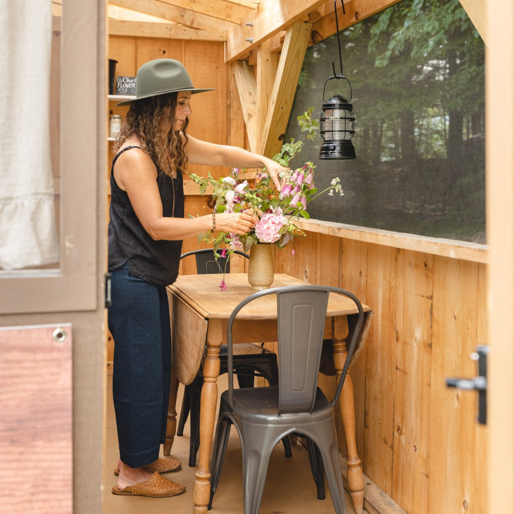 Woman with long curly hair wearing a wide brim sun hat is putting together a floral arrangment on a small dining table inside a wooden cabin