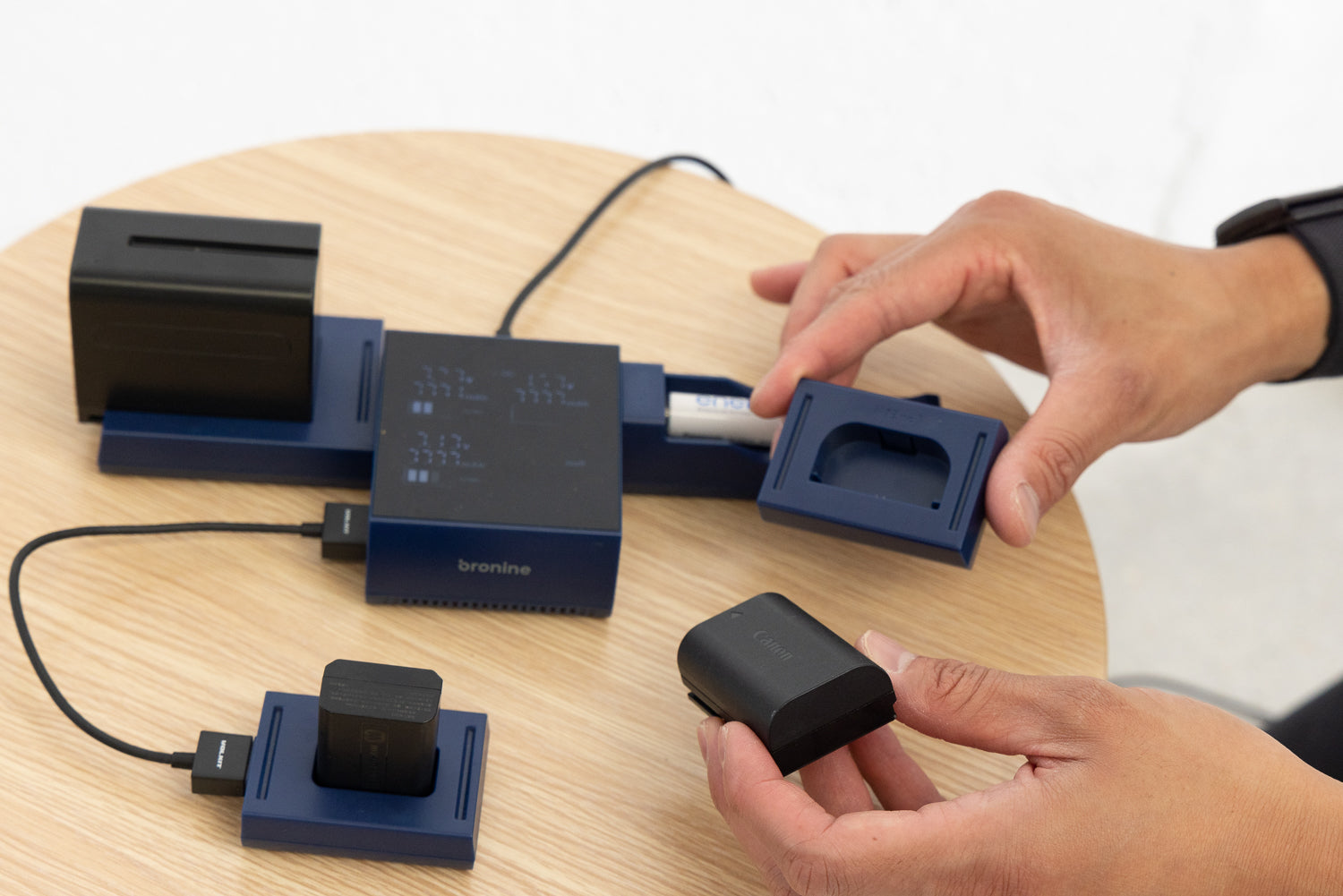 An image of a hand holding a camera battery and plugging it into one of the four ports of a Bronine 4-port multi-charger, which is sitting on a wooden table. The charger is currently active and charging the various camera batteries, with LED lights indicating their progress. Three other camera batteries are also plugged into the charger's remaining ports.