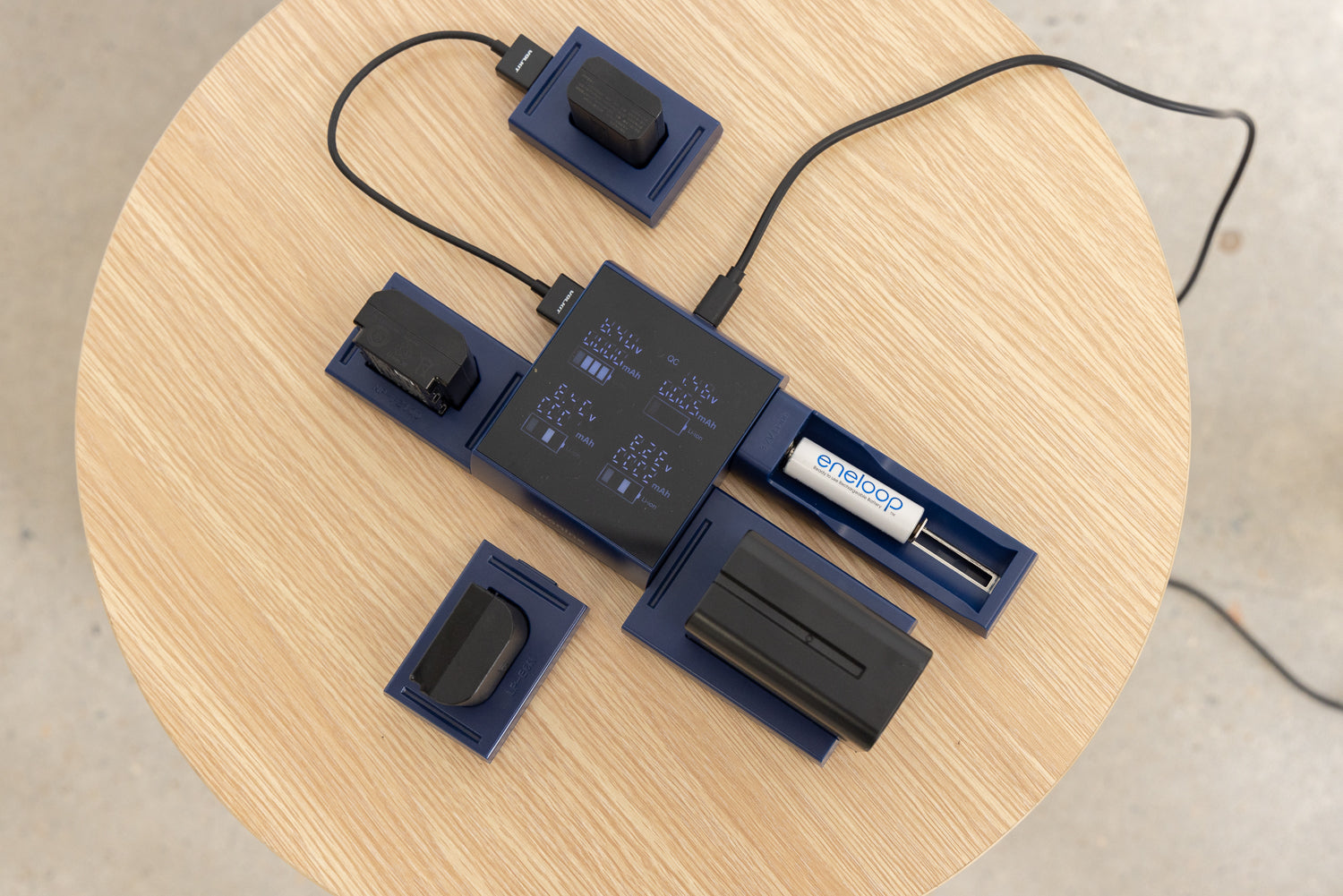 An image of a Bronine 4-port multi-charger sitting on a wooden table, with four different types of camera chargers plugged into its ports. The charger is currently active and charging the various camera batteries.