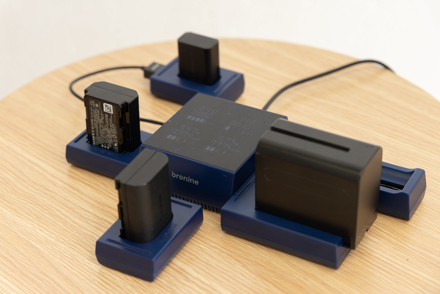 An image of a Bronine 4-port multi-charger sitting on a wooden table, with four different types of camera chargers plugged into its ports. The charger is currently active and charging the various camera batteries.