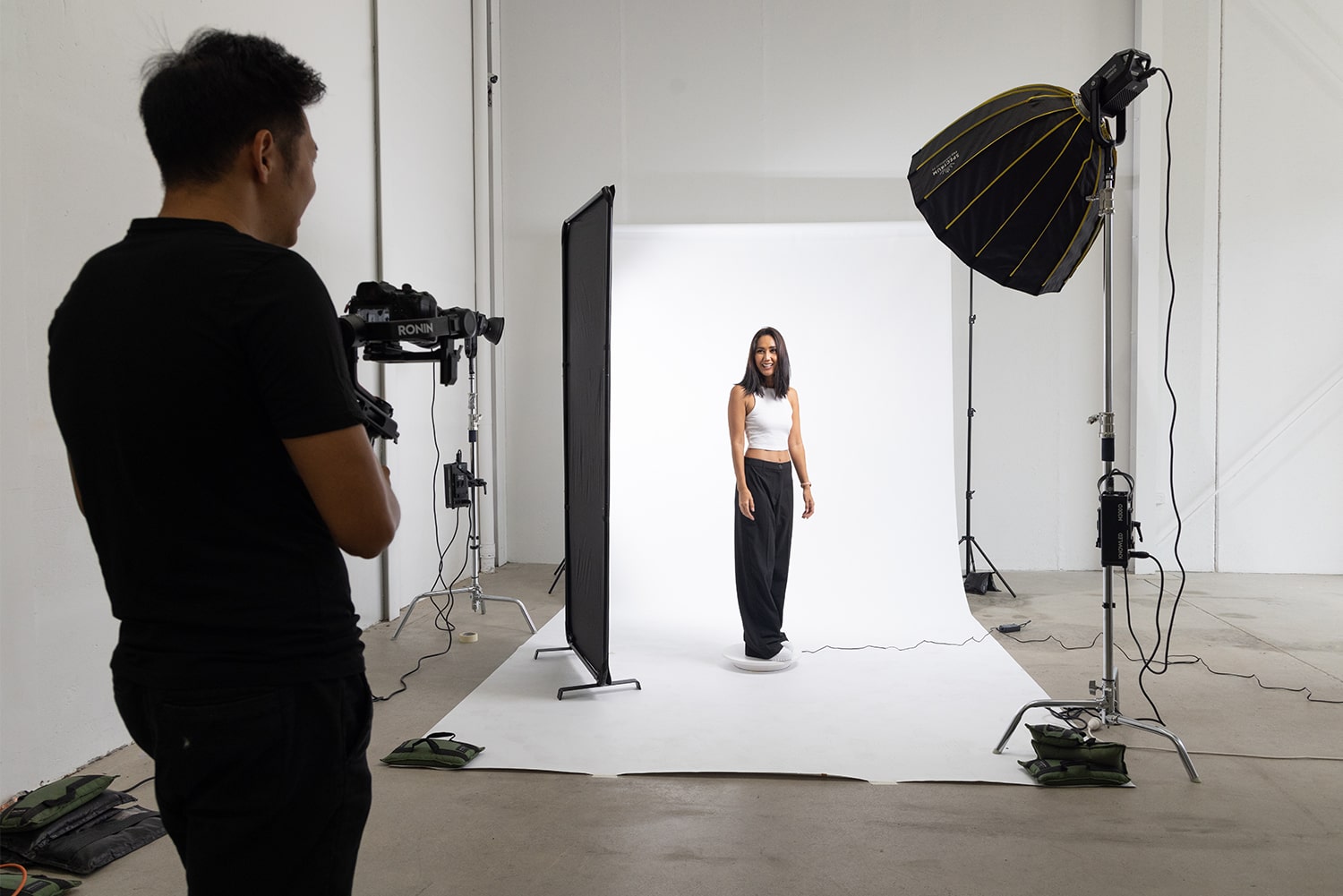 An image of a professional photo studio setup featuring a model standing on a 360-degree turntable, dressed in a white halter top and black pants. The model is smiling and posing confidently while a photographer is taking her photo. The studio is equipped with a seamless white paper backdrop, a v-flat backdrop, and a studio light, creating a well-lit environment for fashion photography.