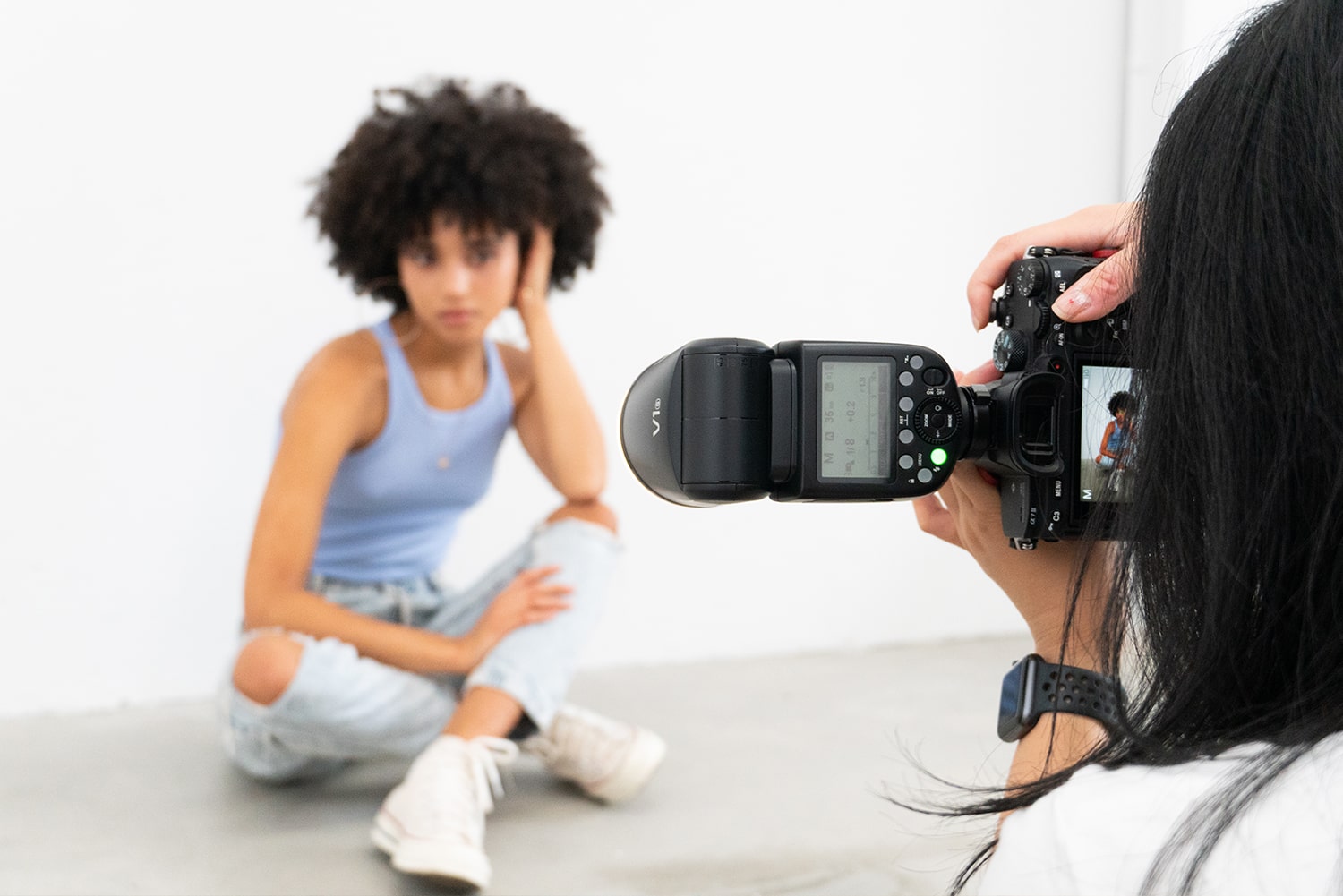 a photographer capturing an image of a curly-haired model who is seated. The main focus of the image is the camera held by the photographer, poised to capture the model's photo.