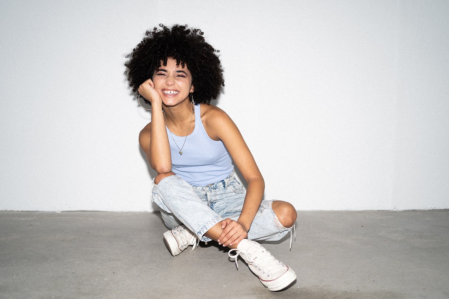 Full length shot of a curly-haired model with a radiant smile, looking directly at the camera. The model is seated in an Indian sit position, also known as a cross-legged sitting position, with one hand resting on her cheeks.