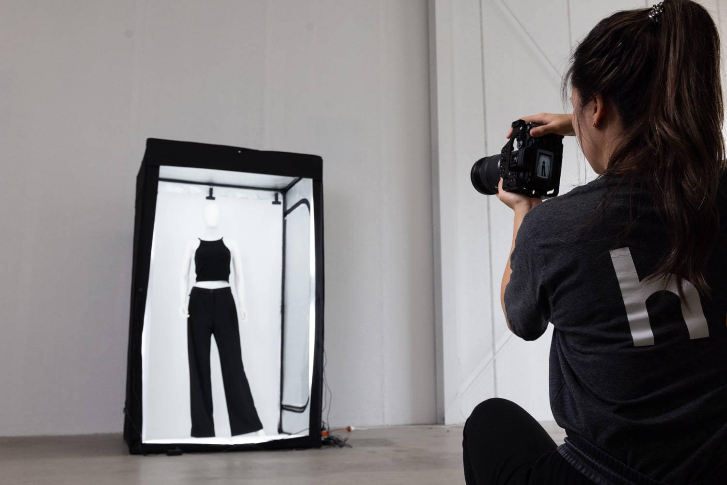 An image showing a photographer taking a picture of a black halter top and black pants displayed on a mannequin, inside a mini studio designed for fashion photography.