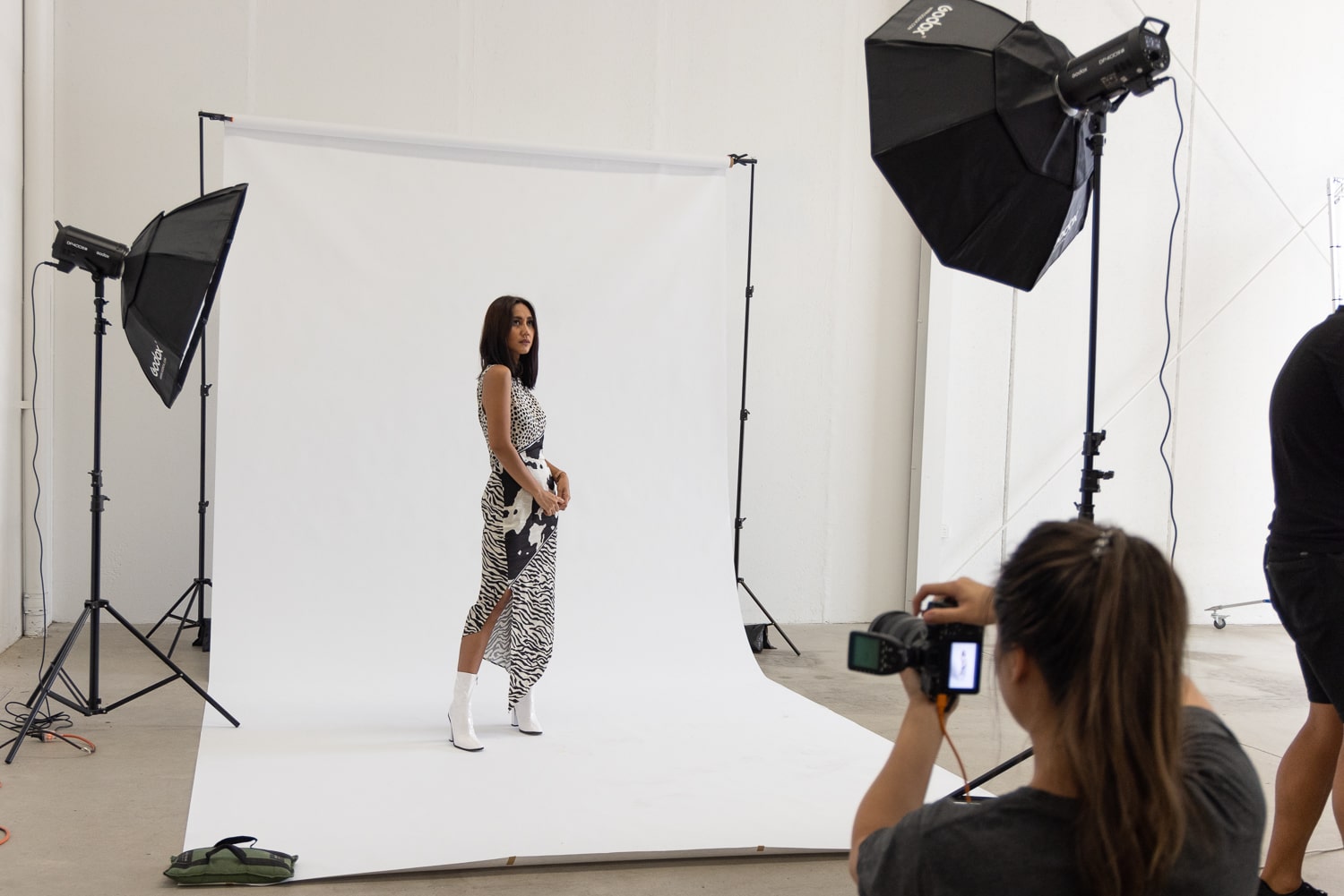 An image of a photographer in a studio setup, capturing a photo of a model dressed in a black and white dress with white boots. The model is posing confidently in front of a white seamless paper backdrop, and the studio is equipped with two lights to illuminate the scene.