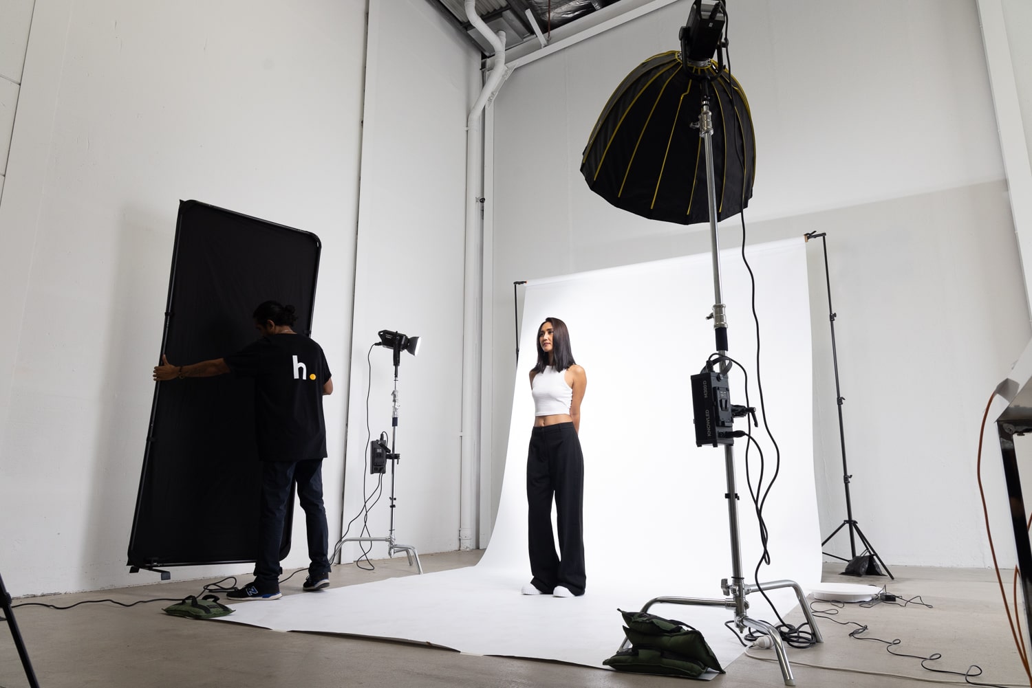An image of a studio setup featuring a model standing on a seamless white paper backdrop, dressed in a white halter top and black pants. Beside the model, another photographer is setting up an Xpress v-flat backdrop. The studio is equipped with two professional lighting equipment, creating a well-lit environment for e-commerce fashion photography.