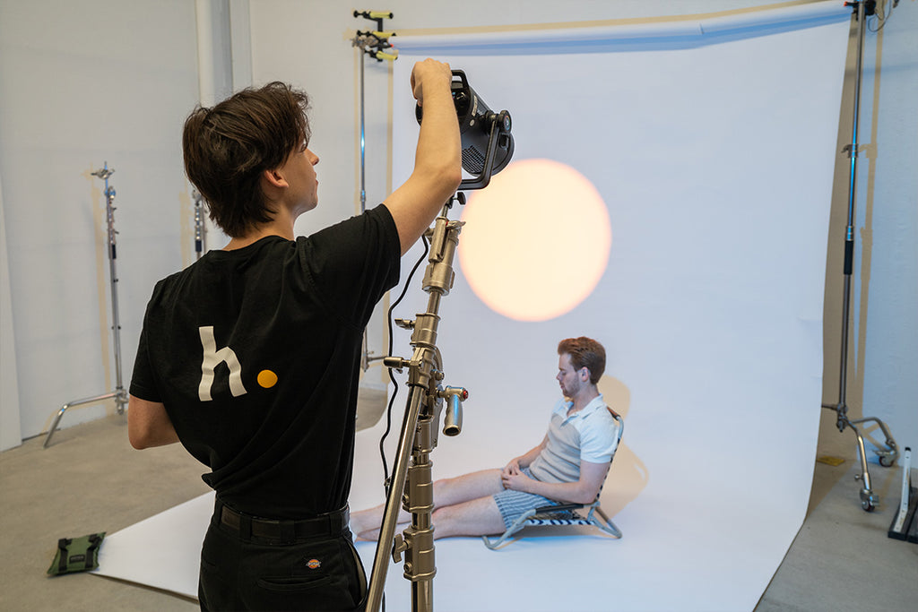 Photographer adjusting a spotlight while a male model sits on a light coloured backdrop