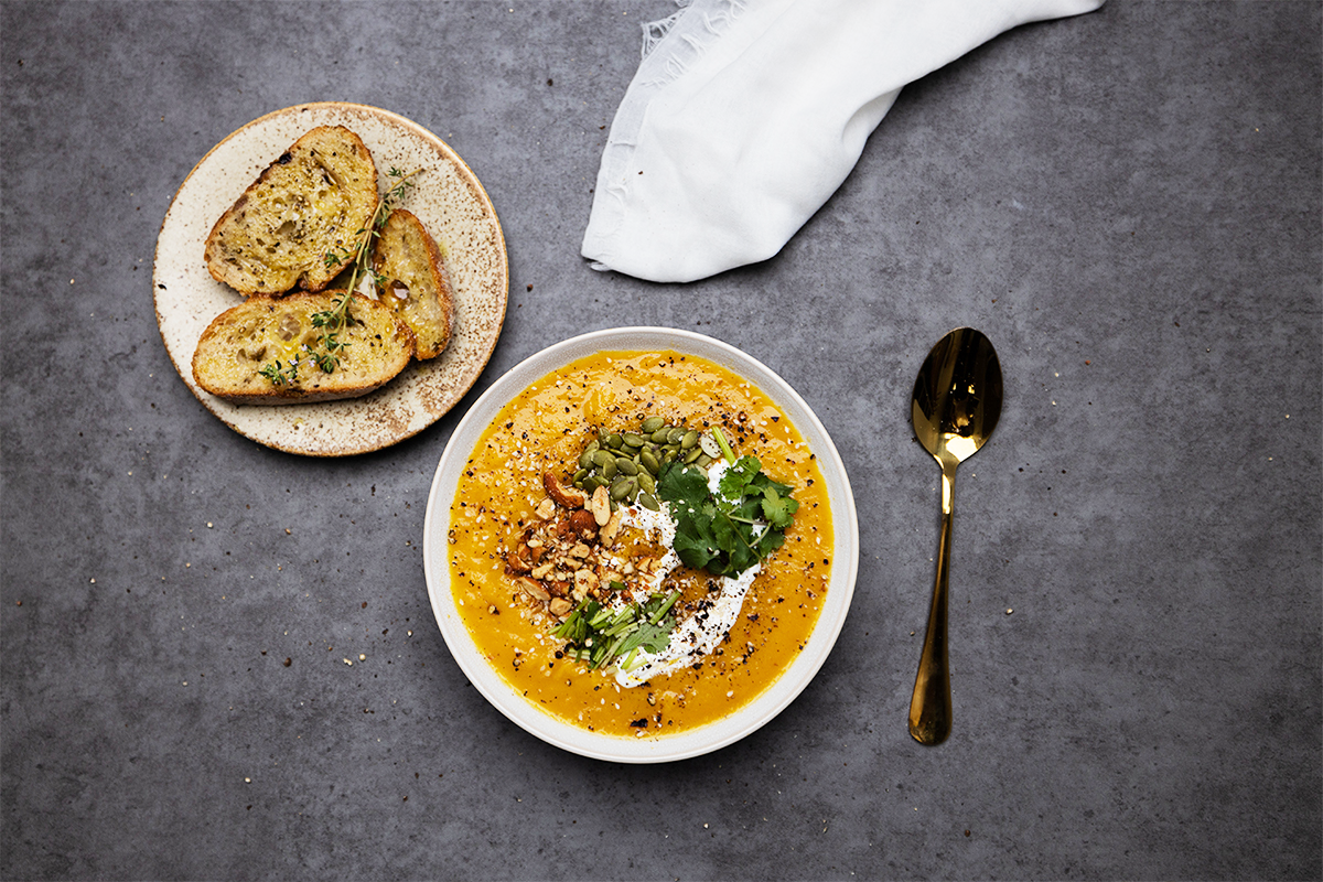 Bowl of pumpkin soup next to a plate of bread, a spoon and white muslin placed on a grey flat lay backdrop