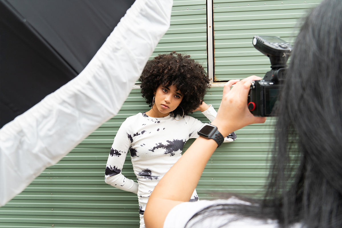Model holding the side of her head in front of green metal while getting her photo taken by a photographer