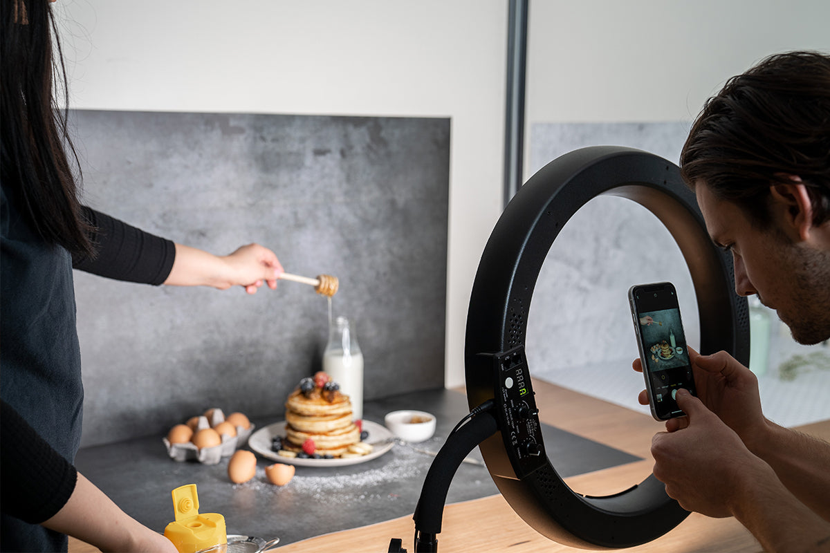 Male photographer using a ring light to take iPhone photos of pancakes and eggs on a grey textured backdrop. Woman drizzles honey over the pancakes