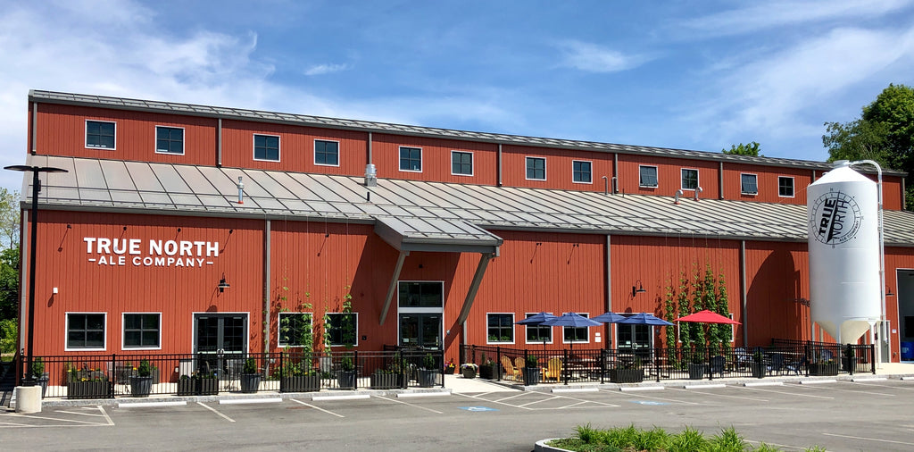 The front of True North Ale Company's brewery and taproom. A large red barn-like building with fenced-in patios, a large white grain silo with the True North logo, and hops growing up the side of the building.