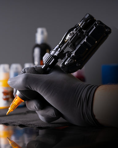 Gloved tattoo artist's hand holding the InkJecta Flite X1 wireless tattoo machine at a tattoo workstation with World Famous Ink in the background