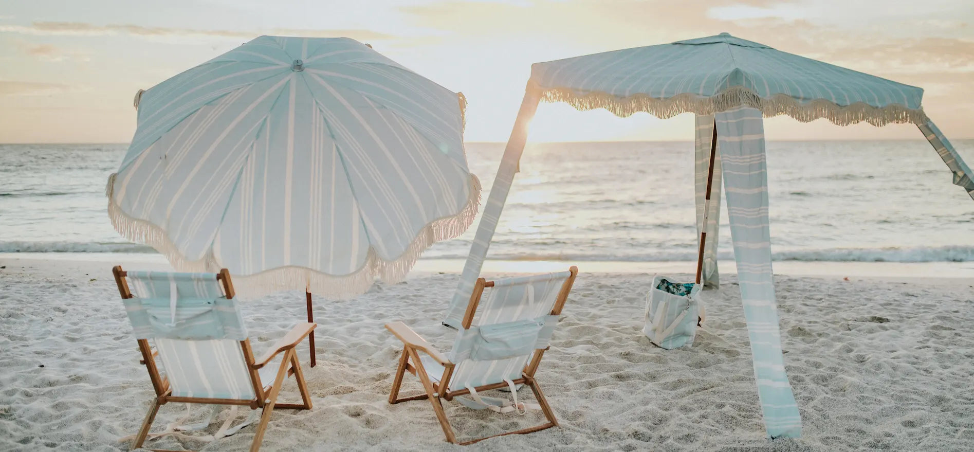 Blue striped umbrella, chair, cooler tote and cabana on the beach during sunset