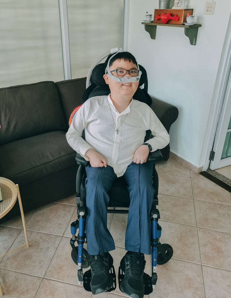 Shalom, a young man, seated in a wheelchair dressed in a customised white shirt and blue long pants