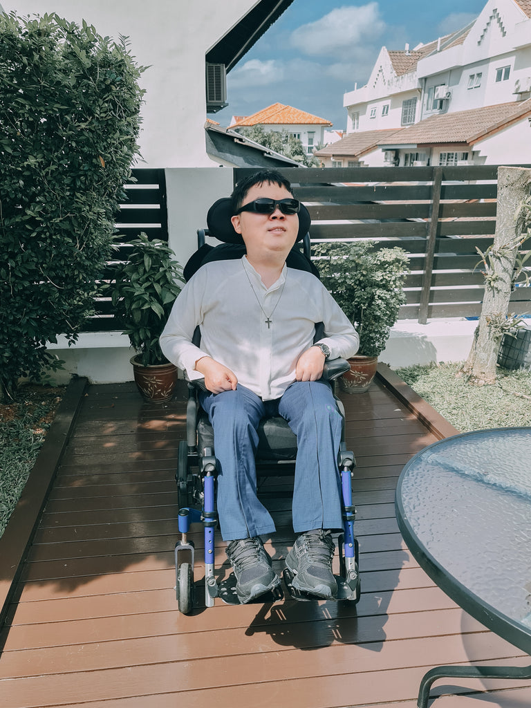 Shalom, a young man, seated outdorrs in his wheelchair while wearing a customised white shirt and blue long pants. He completes the look with a stylish pair of sunglasses.