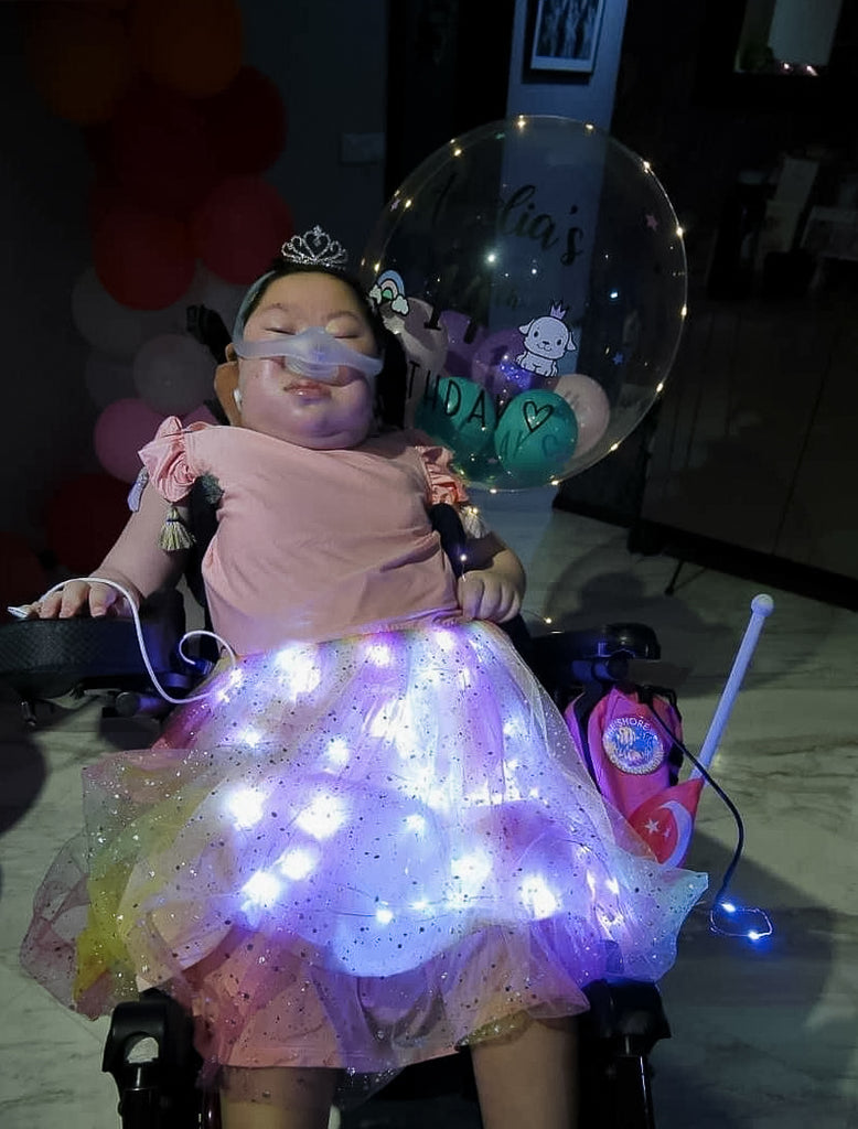 Amelia seated in a wheelchair and wearing her pink dress with lighted up skirt. a 14th birthday balloon is next to her.