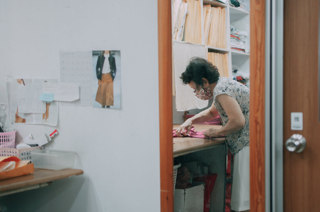 A reflection of an elderly lady in a mirror, hunched over a table as she works on dressmaking.