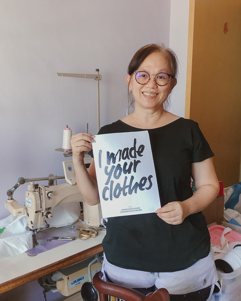 A Chinese lady in her 50's holding up a white A4 sized paper with the text "I made your clothes". She is standing in front of her workstation with a sewing machine in the background.