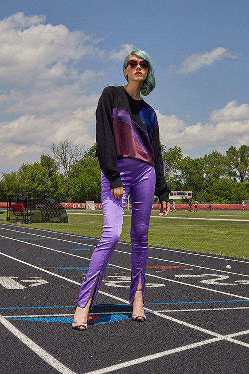 Cynthia Rowley 2019 Resort Collection features a black sweater with a pink, purple, and blue design, worn with silk purple pants that have a slight slit at the bottom of the pant leg.