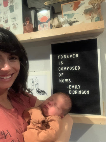 Jonnie holding her baby in front of her art wall. Felt board behind her says "Forever is composed of nows" Emily Dickinson