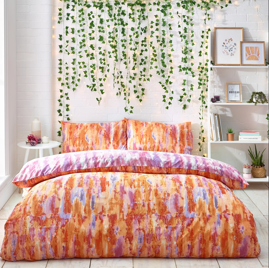 Vibrant bedding in an abstract pattern set with shades of orange, purple and blue, set against a white bedroom with hanging greenery and shelves.