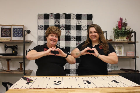 Team members Margo & Kelly making hearts with their hands in front of a quilting display